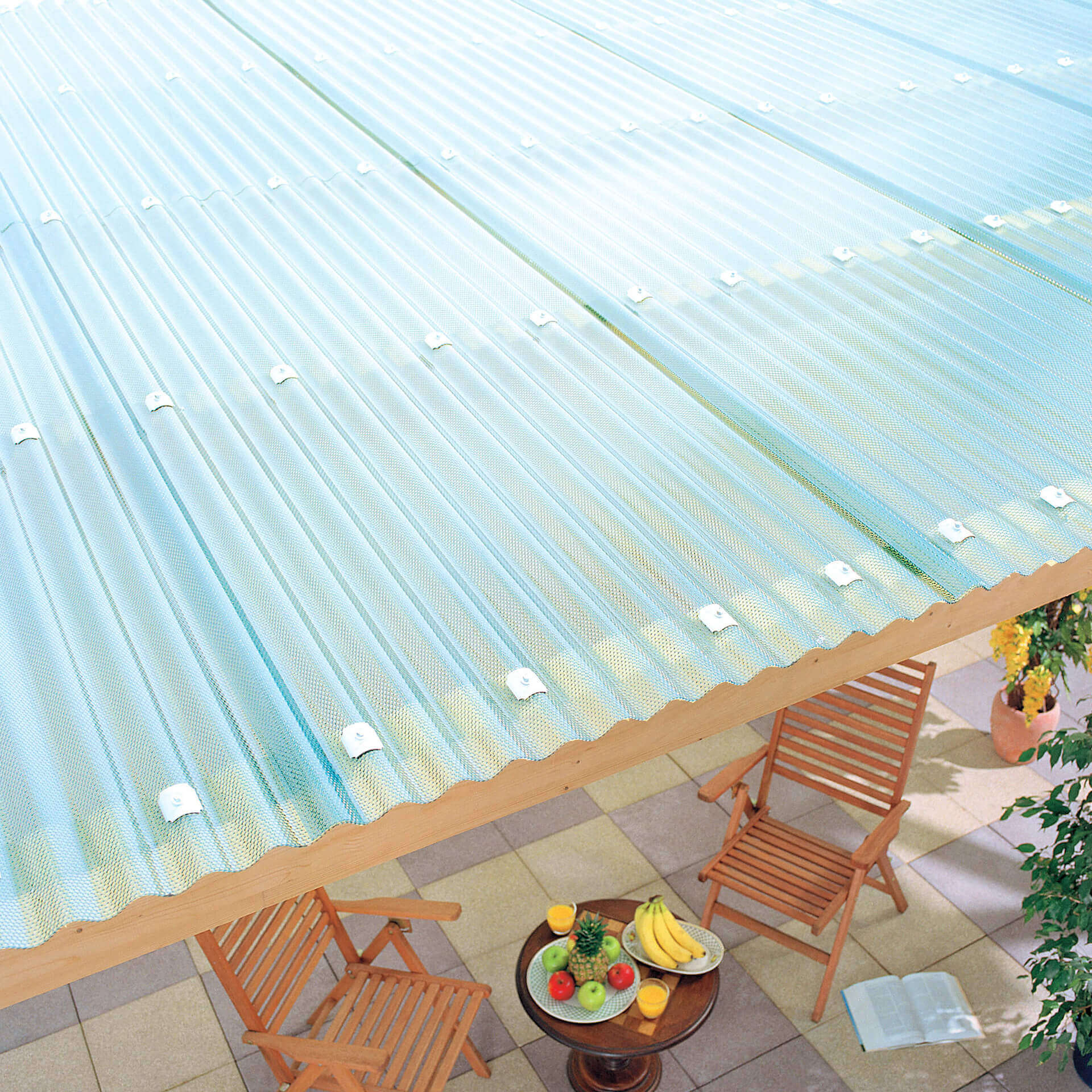 Das Bild zeigt klarbläuliche Wellenplatten, die als Dach über einer Terrasse montiert sind. Darunter stehen Gartenmöbel.